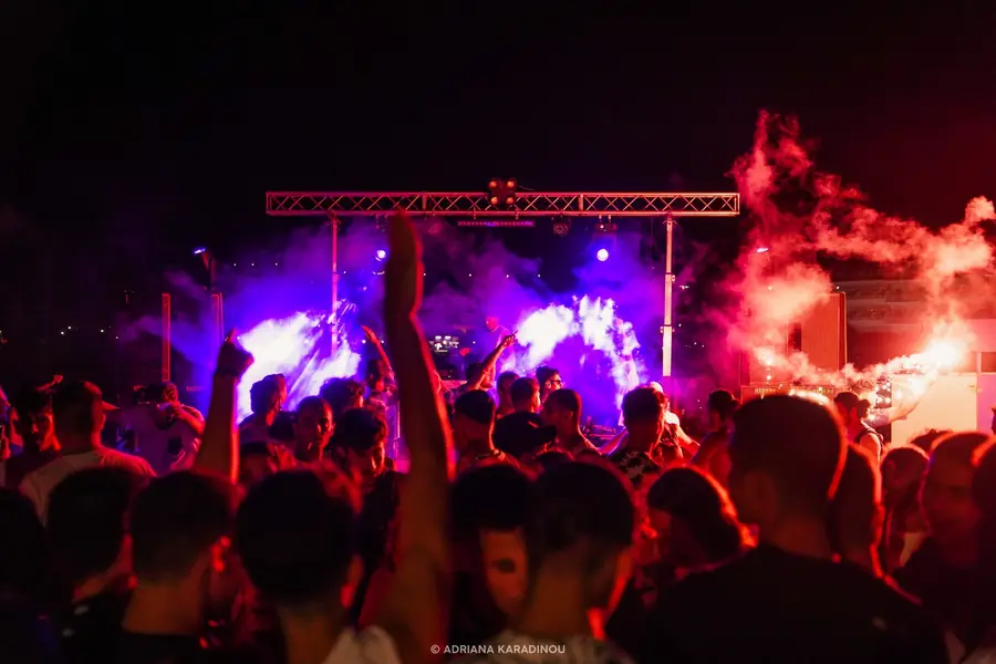 A crowd with raised hands in front of a small stage lit in violet, red flare smoke drifting on the right.
